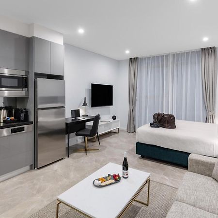 Meriton Suites Sussex Street, Sydney Exteriér fotografie The photo shows a modern studio apartment. On one side, there is a kitchenette with sleek gray cabinets, a refrigerator, and built-in appliances. In the living area, there is a comfortable gray sofa, a white coffee table with a fruit platter and a bo