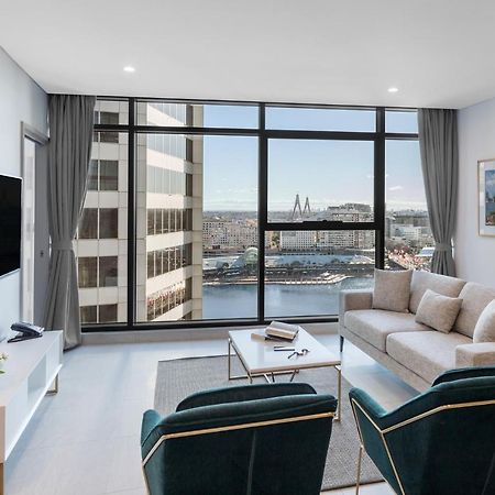 Meriton Suites Sussex Street, Sydney Exteriér fotografie The photo shows a modern living room with large windows that offer a view of the cityscape and water. The interior features a light color palette with pale blue walls and soft lighting. There is a comfortable gray sofa and two green accent chairs, ar