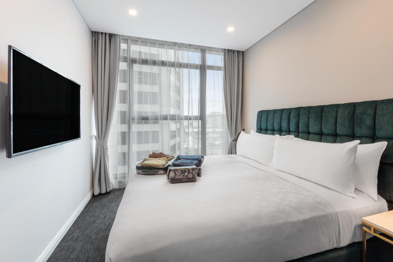 Meriton Suites Sussex Street, Sydney Exteriér fotografie The photo shows a modern hotel bedroom. The room features a large bed with a green upholstered headboard and crisp white linens. There are two pillows on the bed and a few folded items of clothing neatly placed on it. A flat-screen TV is mounted on t