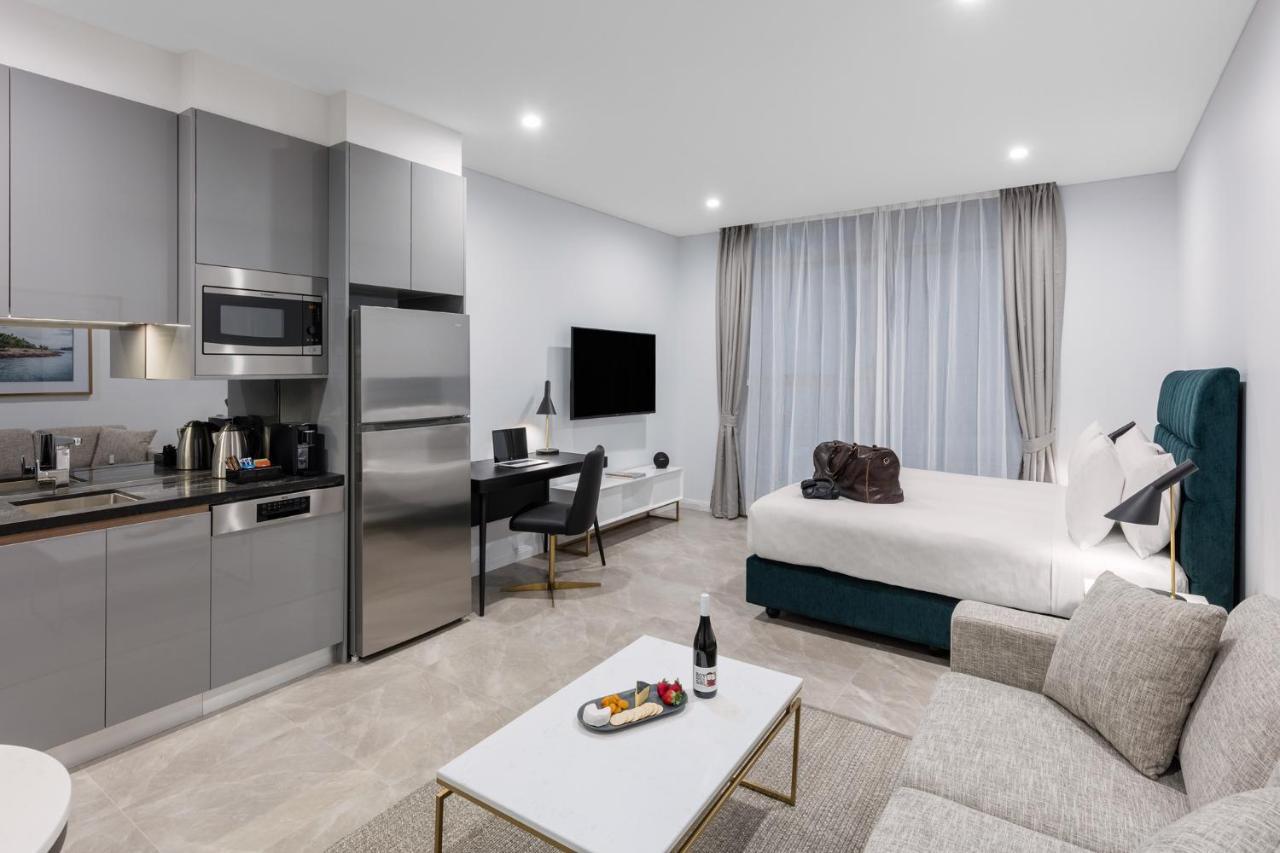 Meriton Suites Sussex Street, Sydney Exteriér fotografie The photo shows a modern studio apartment. On one side, there is a kitchenette with sleek gray cabinets, a refrigerator, and built-in appliances. In the living area, there is a comfortable gray sofa, a white coffee table with a fruit platter and a bo