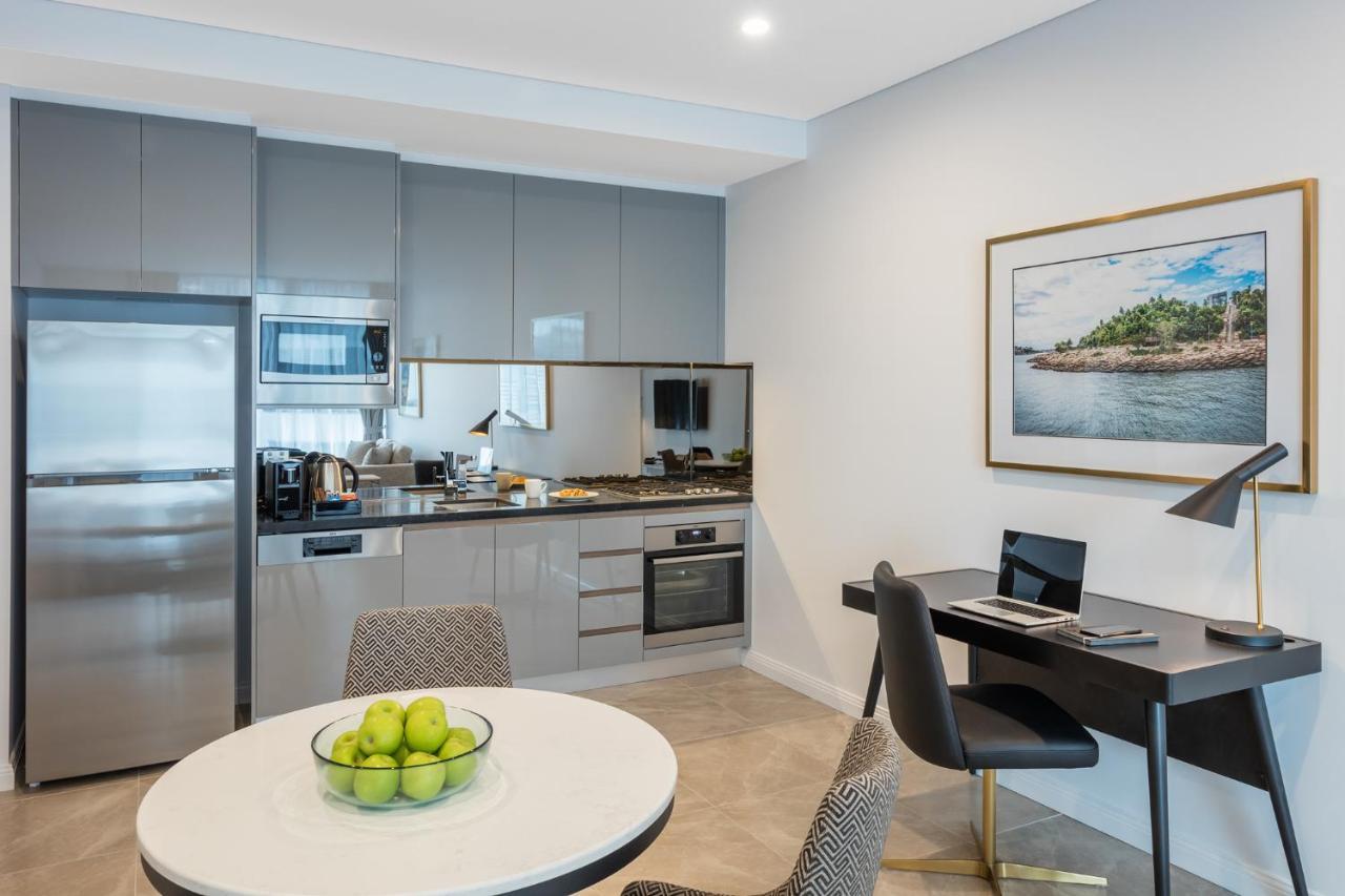 Meriton Suites Sussex Street, Sydney Exteriér fotografie The photo shows a modern kitchen and dining area in an apartment. The kitchen features sleek gray cabinetry, a stainless steel fridge, and a microwave. There is a countertop with cooking appliances, including an oven and stovetop. A small round dinin