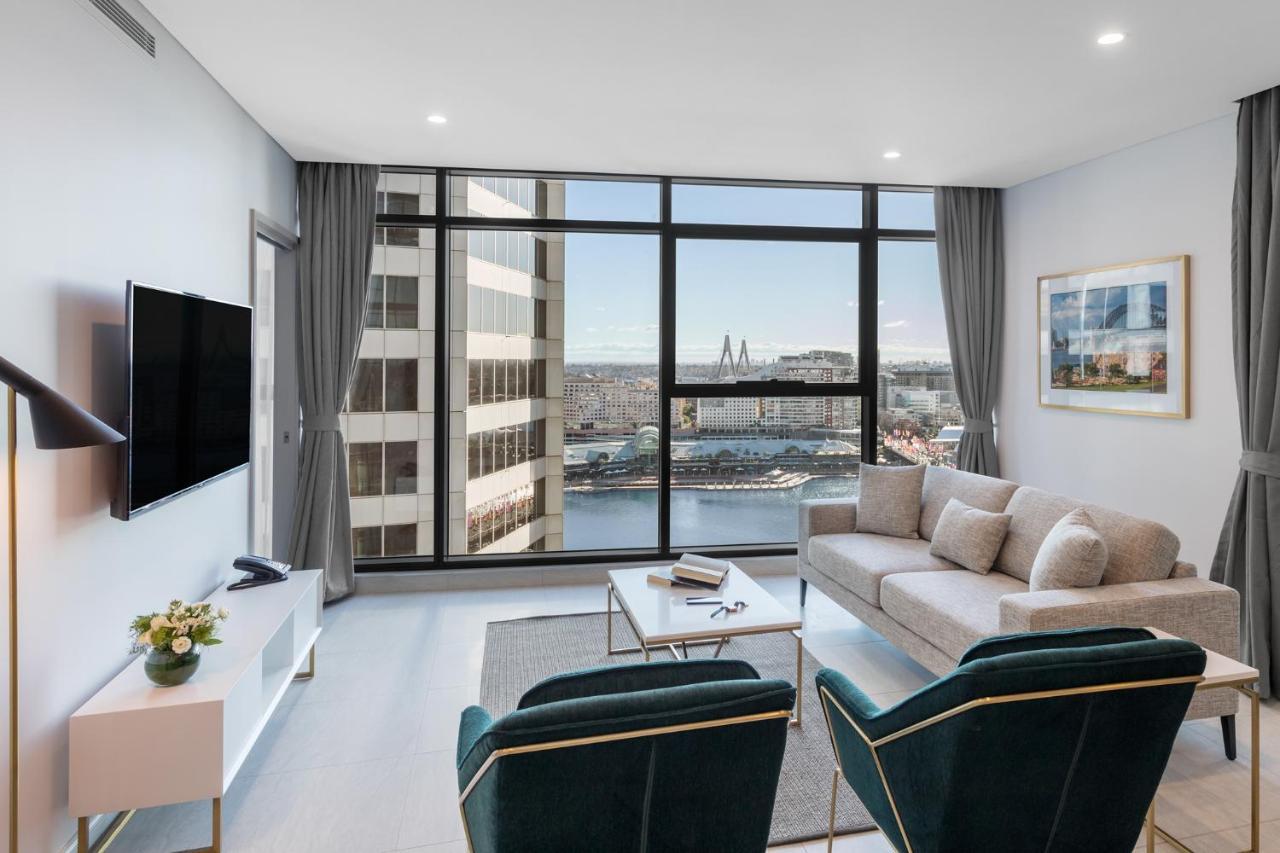 Meriton Suites Sussex Street, Sydney Exteriér fotografie The photo shows a modern living room with large windows that offer a view of the cityscape and water. The interior features a light color palette with pale blue walls and soft lighting. There is a comfortable gray sofa and two green accent chairs, ar