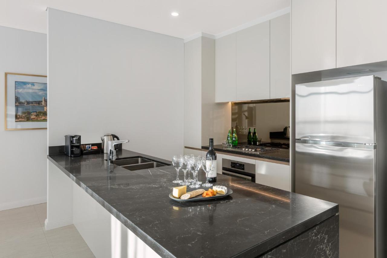Meriton Suites Sussex Street, Sydney Exteriér fotografie The photo shows a modern kitchen featuring a sleek design with a dark stone countertop. There is a built-in sink and possibly a cooktop. The kitchen cabinetry is light-colored, creating a contrast with the dark counter. On the countertop, there are w