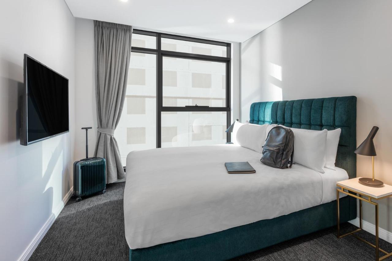 Meriton Suites Sussex Street, Sydney Exteriér fotografie The photo shows a modern hotel room featuring a comfortable bed with white bedding and a teal-colored headboard. There is a neatly placed black backpack on the bed, along with a small notebook or tablet. A television is mounted on the wall opposite t