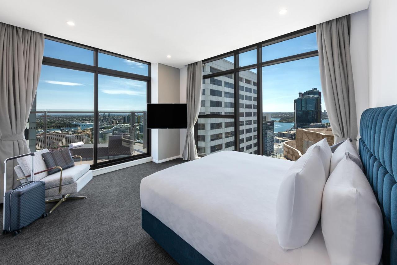 Meriton Suites Sussex Street, Sydney Exteriér fotografie The photo shows a modern hotel room featuring large windows that provide a panoramic view of the cityscape and waterfront. The room is furnished with a king-size bed dressed in white linens, a comfortable armchair, and a flat-screen TV mounted on the