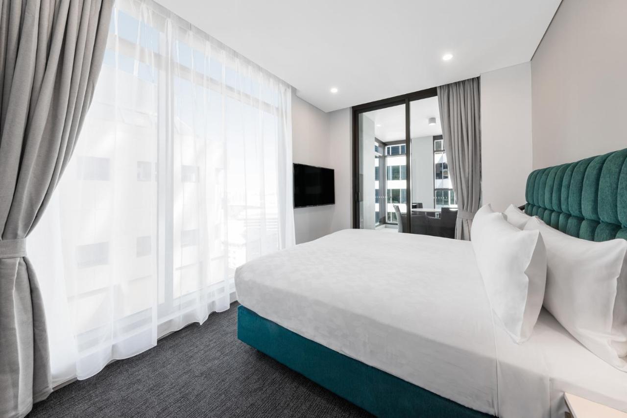 Meriton Suites Sussex Street, Sydney Exteriér fotografie The photo shows a modern bedroom featuring a large bed with a white duvet and pillows. The headboard is turquoise, adding a pop of color to the neutral-toned room. There are sheer white curtains covering large windows that let in natural light, creat