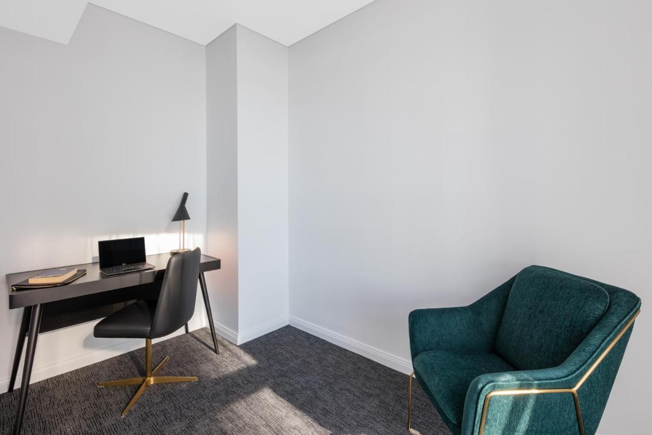 Meriton Suites Sussex Street, Sydney Exteriér fotografie The photo depicts a minimalist interior space, likely a small office or study area. It features a simple, modern desk with a black lamp and a laptop or tablet on it. A sleek black chair accompanies the desk, while on the opposite side of the room, th