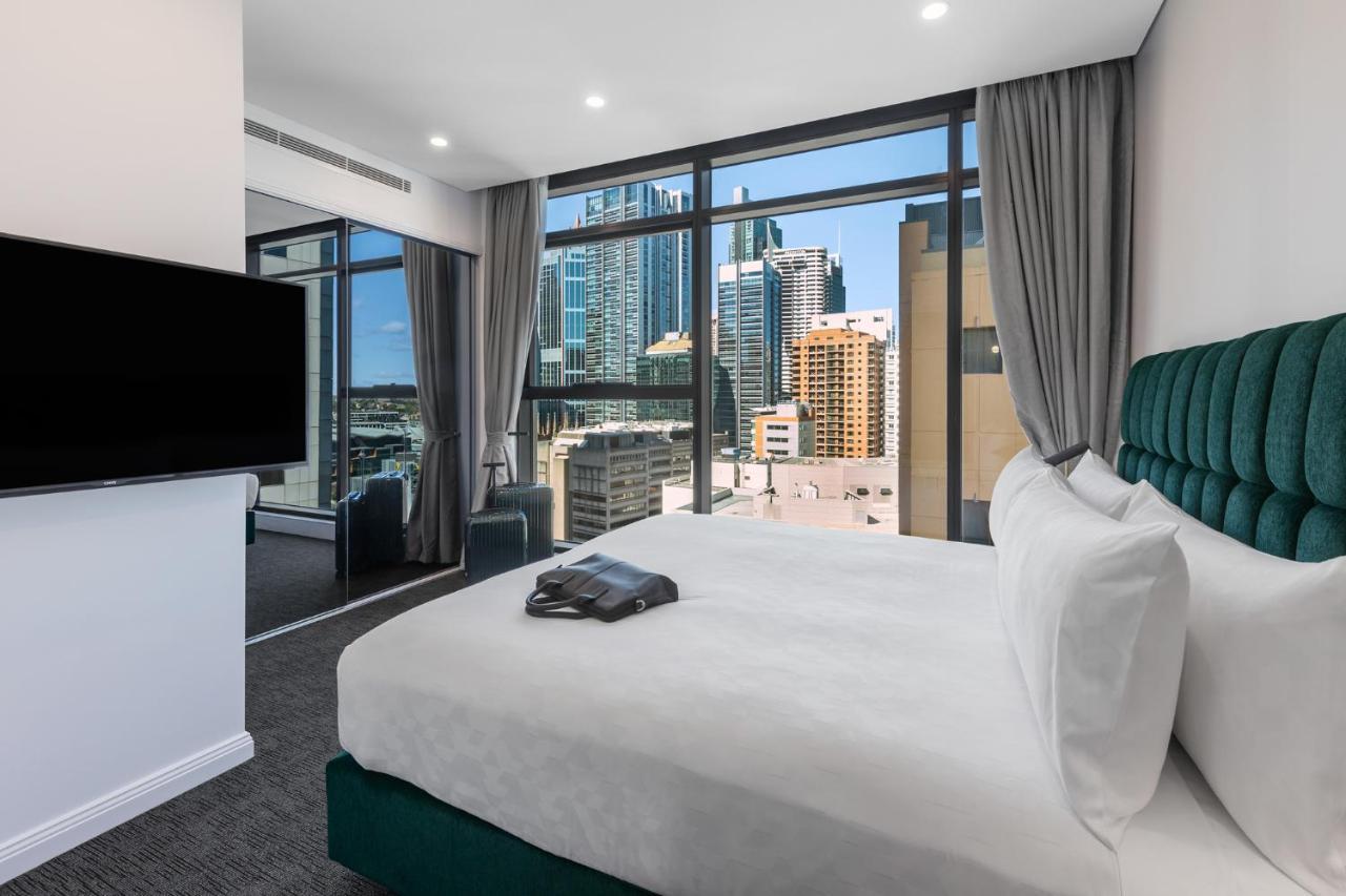 Meriton Suites Sussex Street, Sydney Exteriér fotografie The photo shows a modern hotel room featuring a large bed with white linens and a green upholstered headboard. There is a sleek television mounted on the wall opposite the bed, and the room has large windows that provide a view of a cityscape, includ