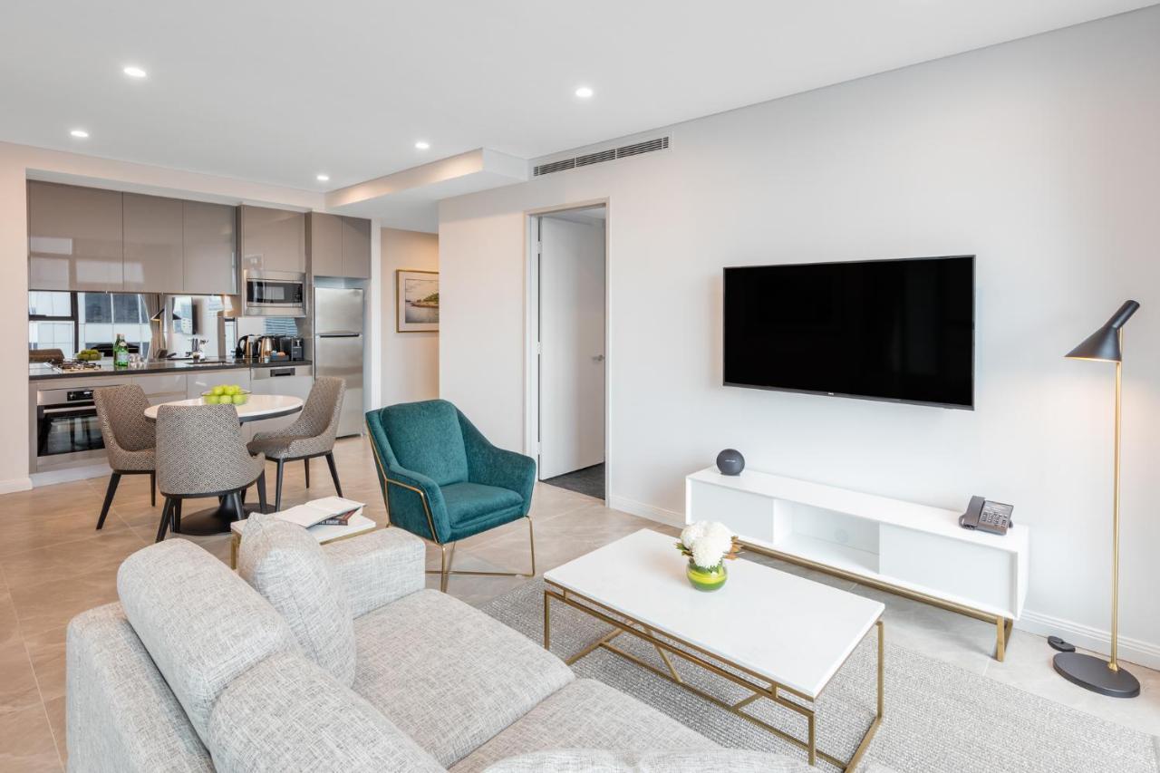 Meriton Suites Sussex Street, Sydney Exteriér fotografie The photo shows a modern living room area. It features a light gray sofa and a coffee table with a decorative plant. There are two chairs, one in teal and a round one, positioned around the coffee table. A flat-screen TV is mounted on the wall, and t