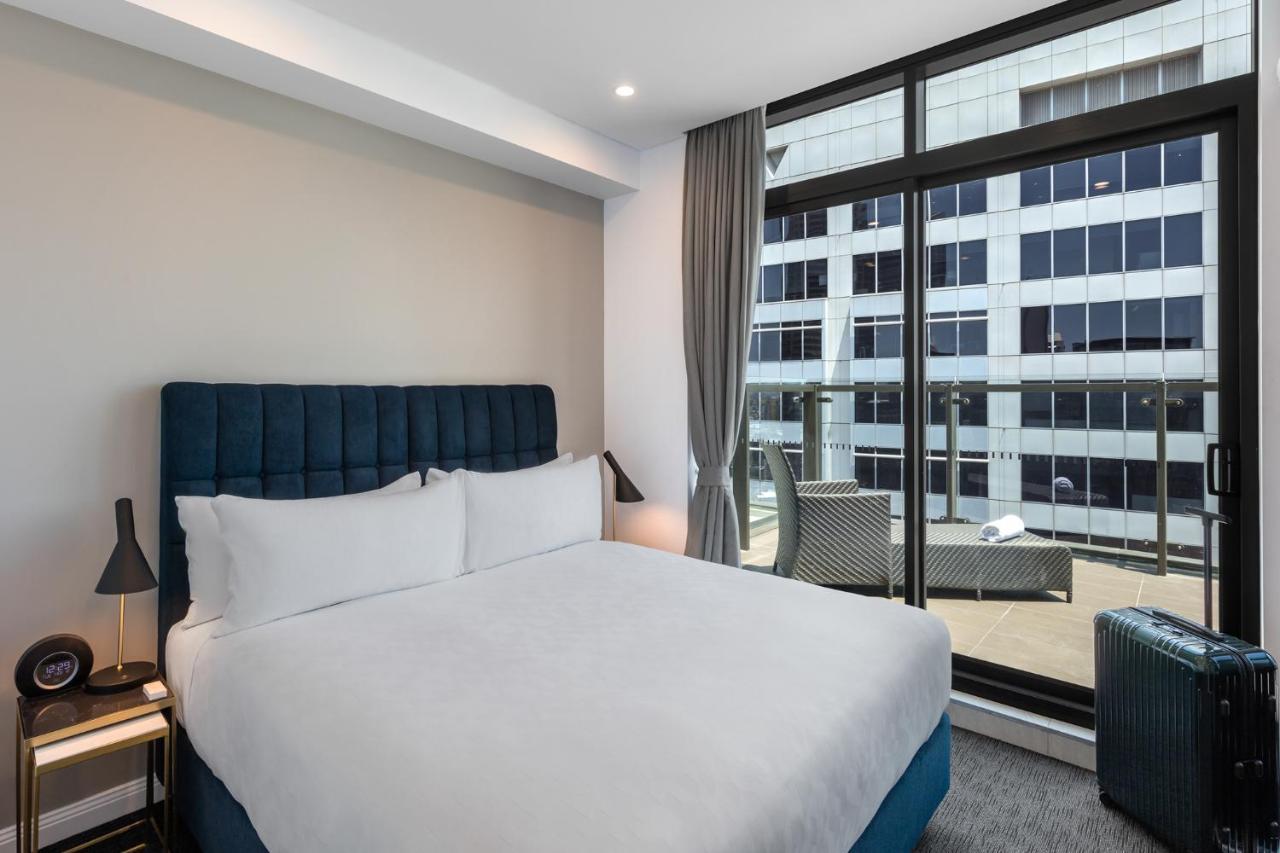 Meriton Suites Sussex Street, Sydney Exteriér fotografie The photo shows a modern hotel room with a large bed featuring a blue upholstered headboard and white linens. Natural light enters through a large window, revealing a view of a building exterior, likely part of the hotel or surrounding area. The room