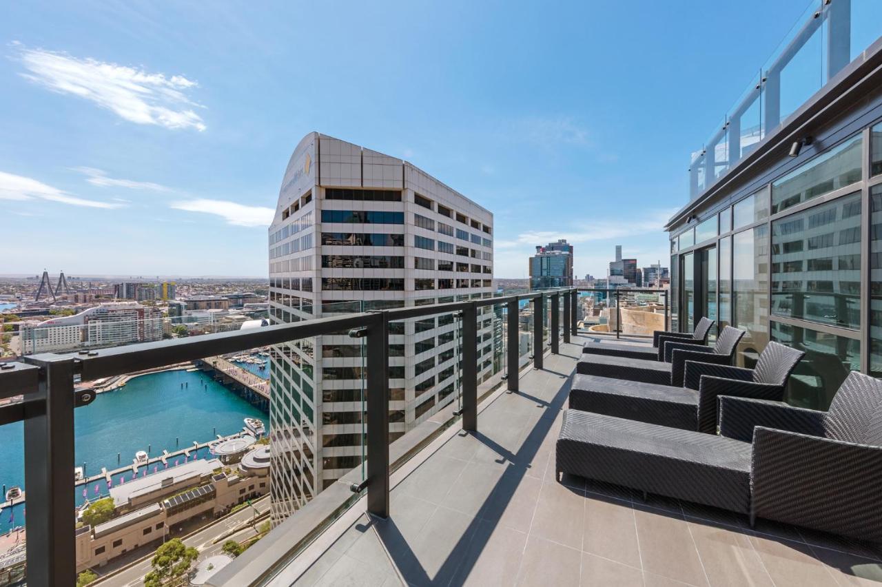 Meriton Suites Sussex Street, Sydney Exteriér fotografie The photo shows a balcony or terrace with a panoramic view of a city skyline. The balcony features modern outdoor furniture, including sleek lounge chairs. In the background, you can see a waterfront area with buildings and a harbor, along with a cle