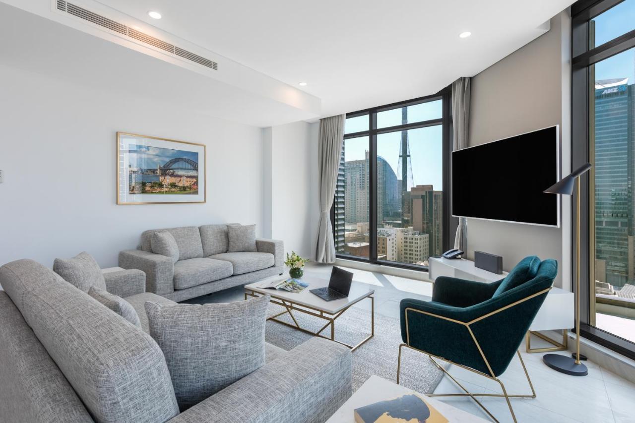 Meriton Suites Sussex Street, Sydney Exteriér fotografie The photo shows a modern living room with large windows offering a view of a city skyline. The room features two gray sofas and a stylish green accent chair. A sleek coffee table is placed in front of the sofas, alongside a laptop. There’s a flat-scr