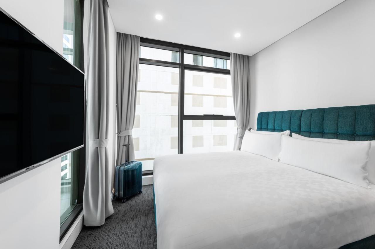 Meriton Suites Sussex Street, Sydney Exteriér fotografie The photo shows a modern hotel room featuring a large bed with a light-colored duvet and a blue upholstered headboard. There are two pillows on the bed. A large window allows natural light to enter the room, and gray curtains frame the window. Next t