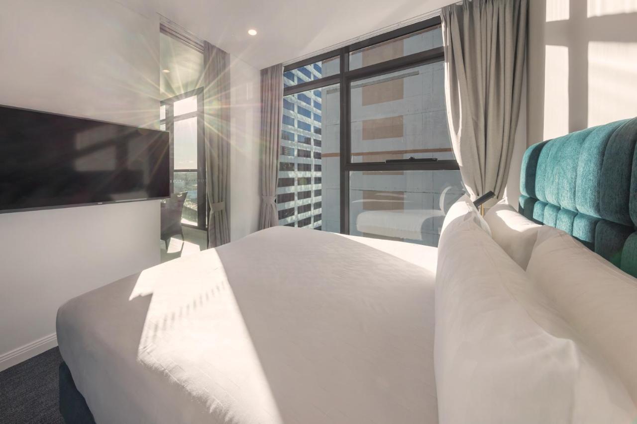 Meriton Suites Sussex Street, Sydney Exteriér fotografie The photo shows a modern hotel room with a neatly made bed featuring white bedding and a plush, tufted headboard in a shade of blue. Natural light is streaming through large windows, which are covered partially by light gray curtains. A flat-screen t