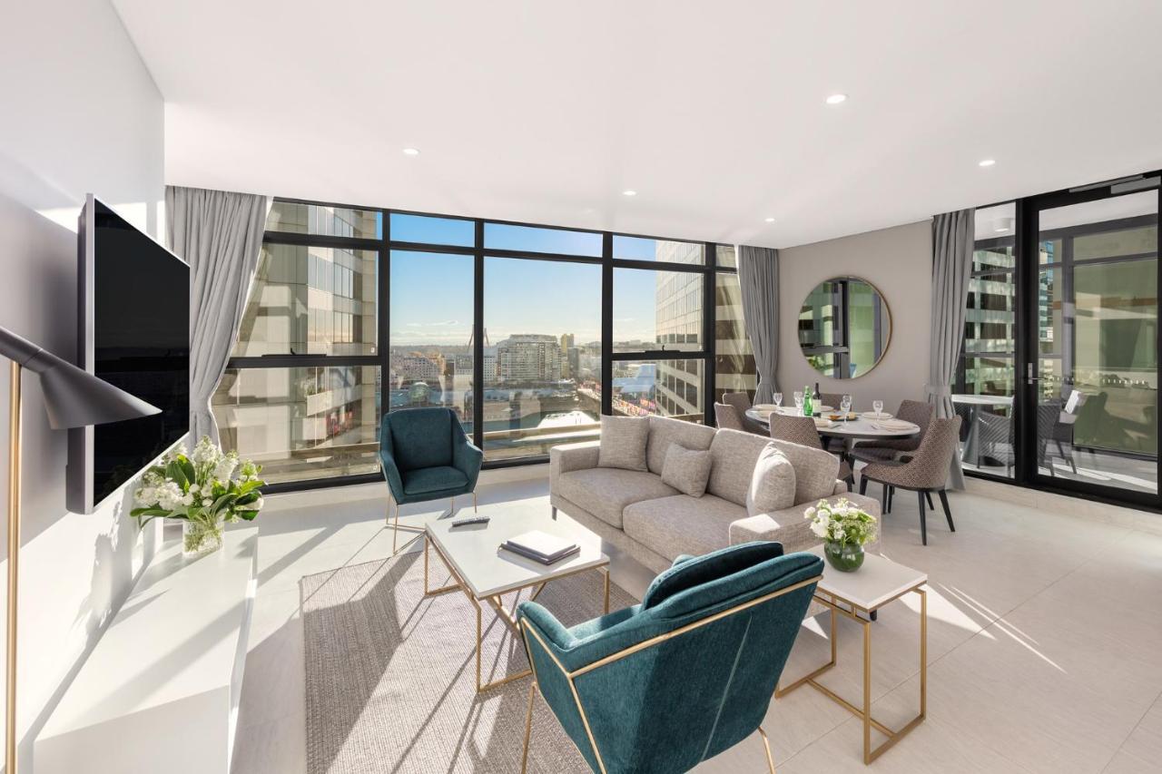 Meriton Suites Sussex Street, Sydney Exteriér fotografie The photo shows a modern and stylish living space. It features a spacious room with large windows that provide a bright view of the cityscape. The furniture includes a light gray sofa, two teal chairs, and a coffee table. There is a flat-screen TV mo