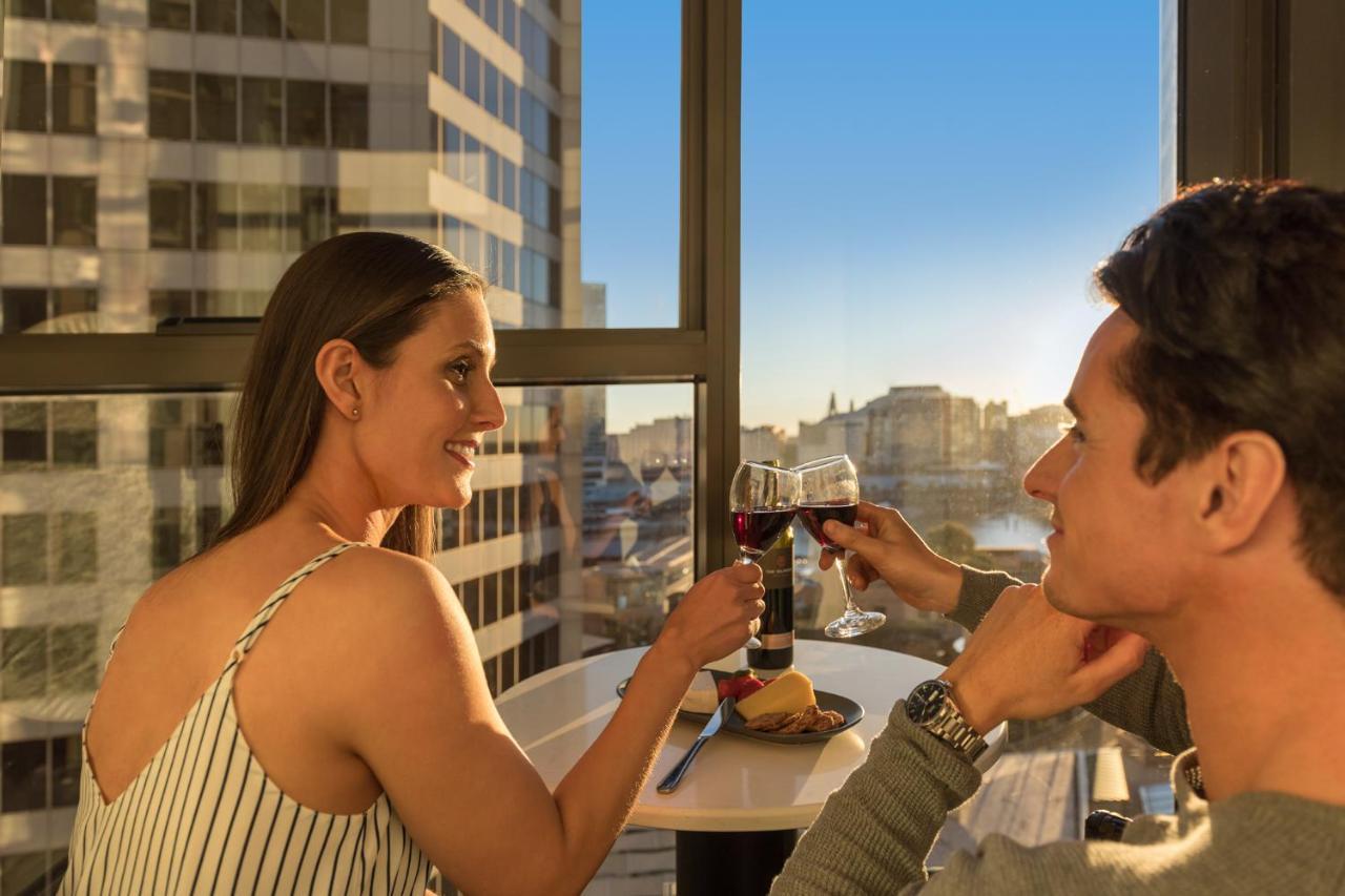 Meriton Suites Sussex Street, Sydney Exteriér fotografie The photo depicts a couple enjoying a drink together in a modern setting. They are sitting at a small table near a large window, which provides a view of a cityscape and a sunset. Both individuals are smiling and engaged in a cheerful toast with glas