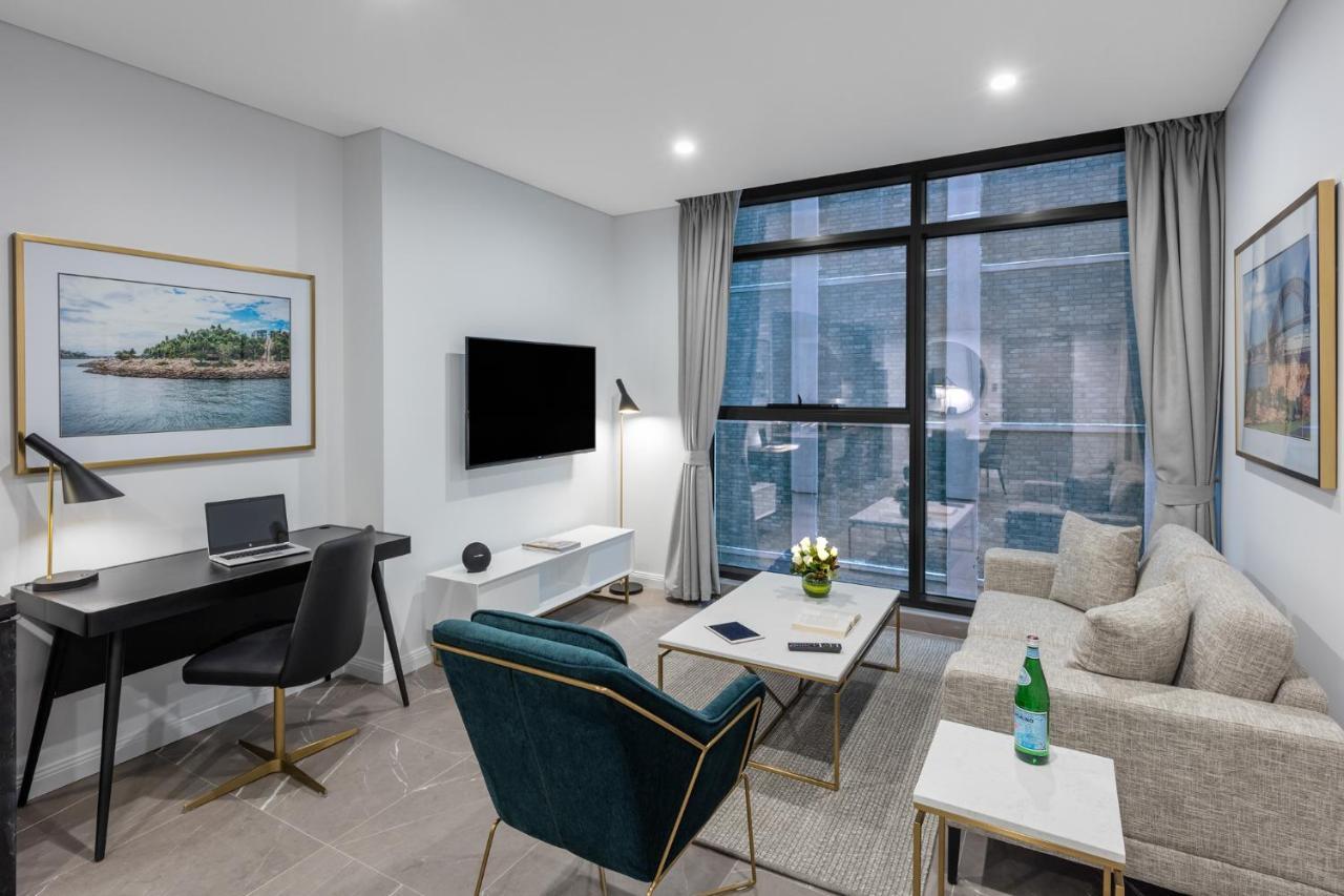 Meriton Suites Sussex Street, Sydney Exteriér fotografie The photo shows a modern living area with a light color scheme. There is a comfortable seating arrangement that includes a beige sofa and an accent chair in teal. A coffee table is positioned in front of the sofa, with a small vase of flowers on it. 
