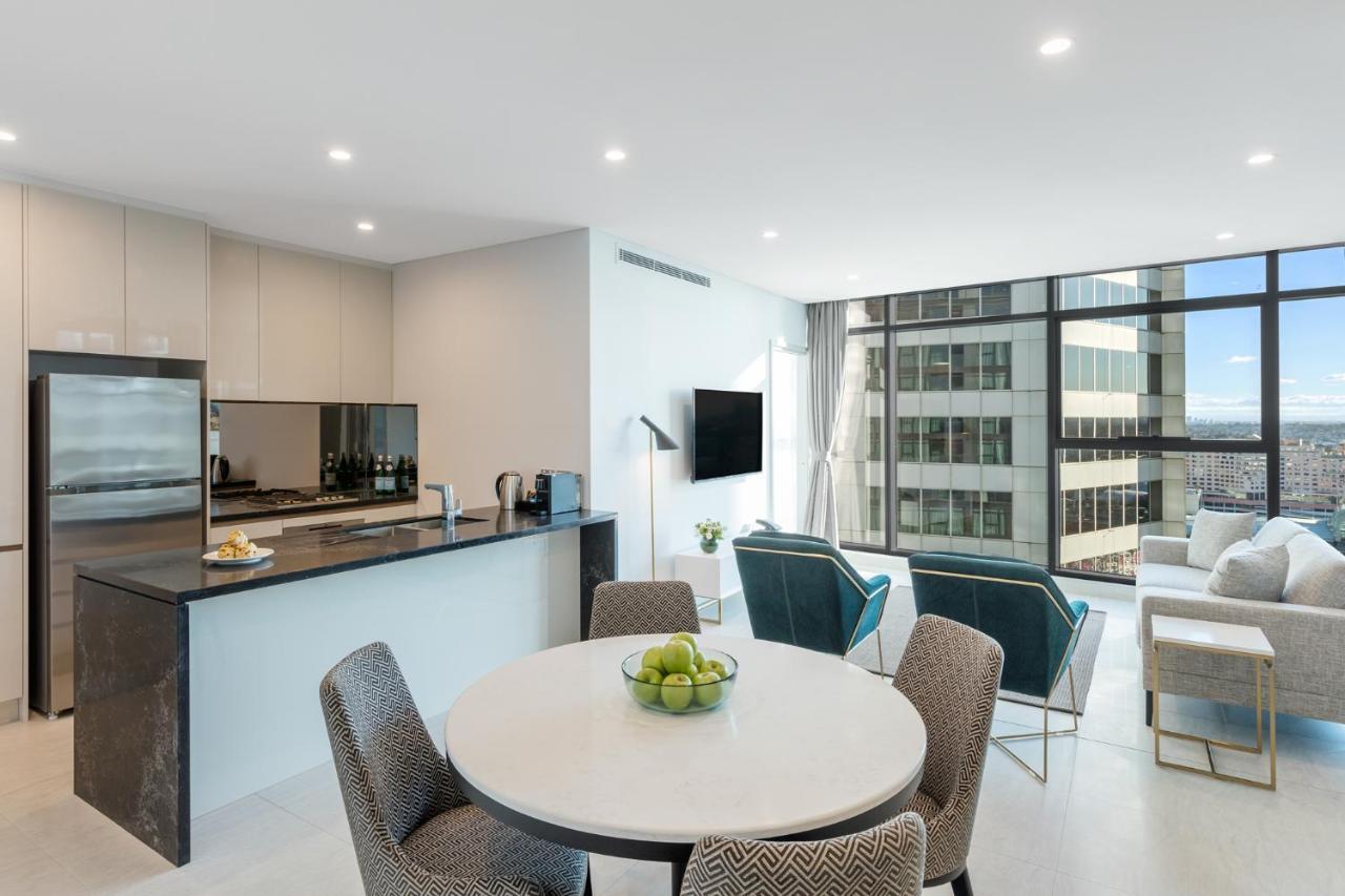 Meriton Suites Sussex Street, Sydney Exteriér fotografie The photo shows a modern and stylish interior space, likely a living room and kitchen area. The kitchen features sleek cabinetry and a countertop, with appliances visible, including a coffee maker. There is a round dining table with gray chairs, set 