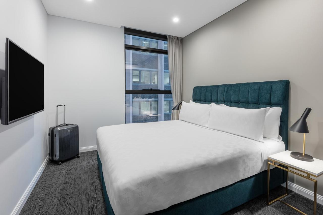 Meriton Suites Sussex Street, Sydney Exteriér fotografie The photo shows a modern hotel room with a clean and minimalist design. There is a large, neatly made bed with white bedding and a teal-colored headboard. On one side of the bed, there is a small nightstand with a lamp. A sleek television is mounted 