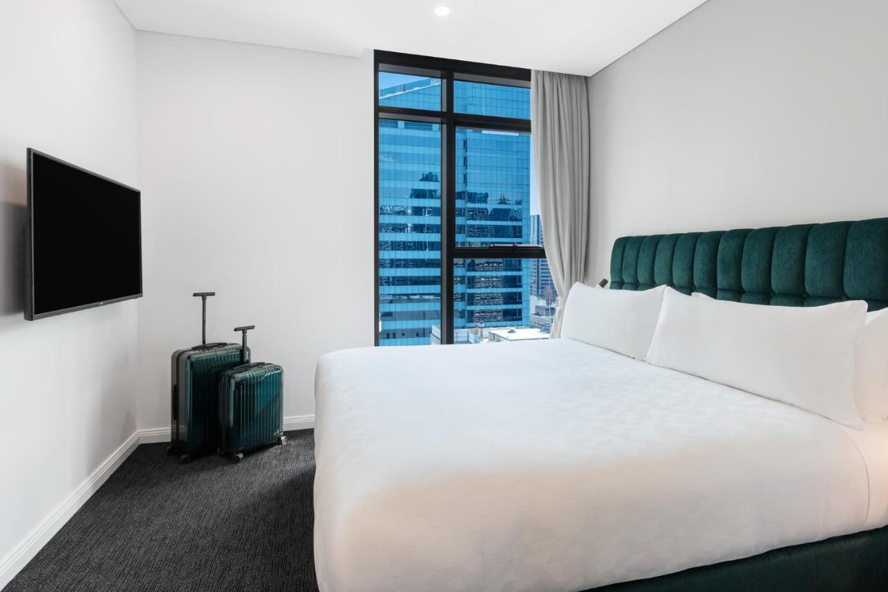 Meriton Suites Sussex Street, Sydney Exteriér fotografie The photo shows a modern hotel room featuring a large bed with white bedding and a green upholstered headboard. There are two green suitcases placed beside the bed. A flat-screen TV is mounted on the wall opposite the bed. The room has a large window