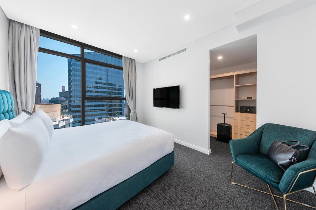 Meriton Suites Sussex Street, Sydney Exteriér fotografie The photo shows a modern hotel room. It features a large window that allows natural light to fill the space, offering a view of the city skyline. The room includes a neatly made bed with a white duvet and a teal-colored base. There is a small green a