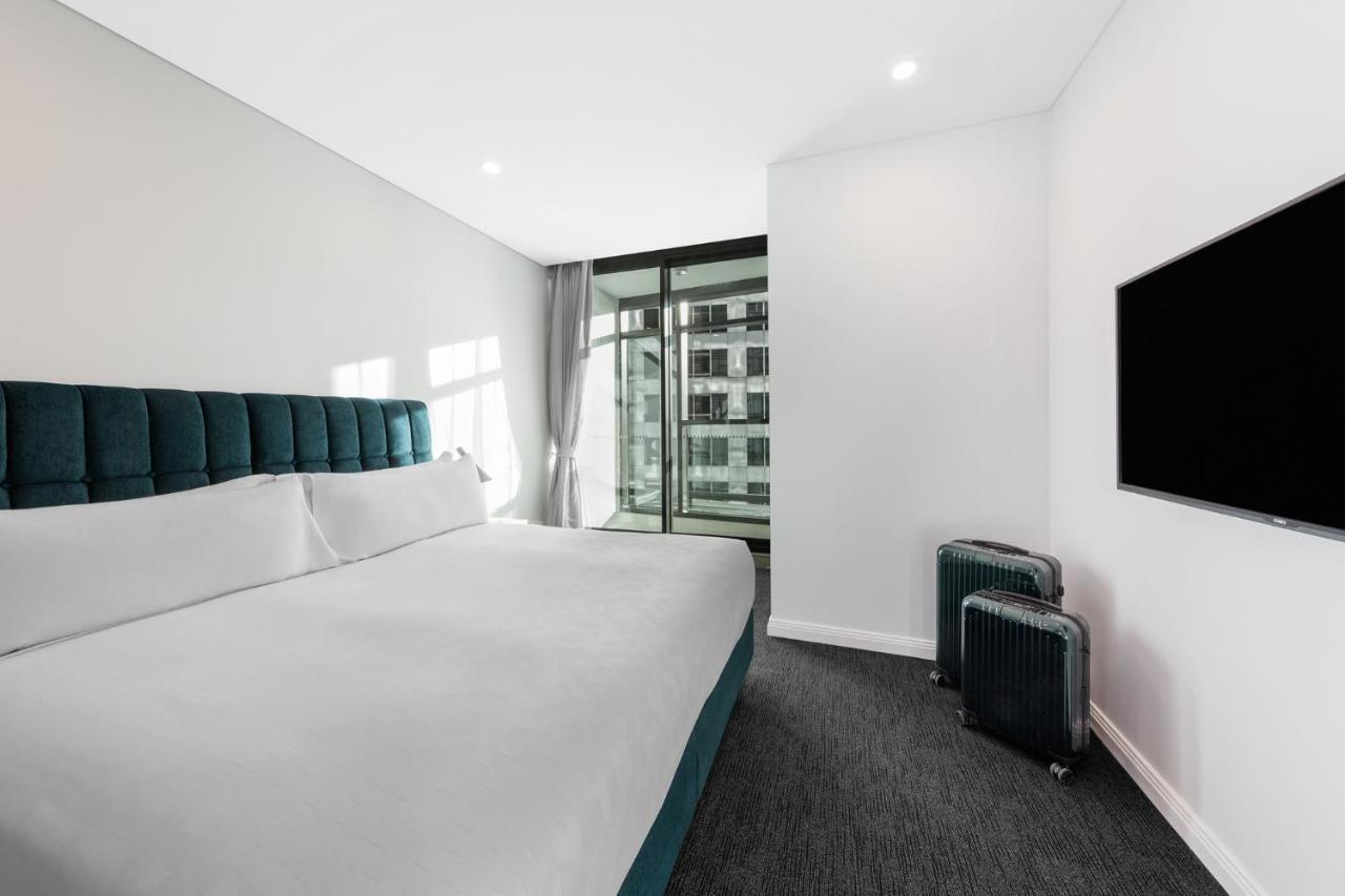Meriton Suites Sussex Street, Sydney Exteriér fotografie The photo shows a modern hotel room featuring a large bed with a headboard in a teal color. The bedding is white and appears neatly arranged. There is a large window allowing natural light to enter, offering a view of an outdoor area. A flat-screen T