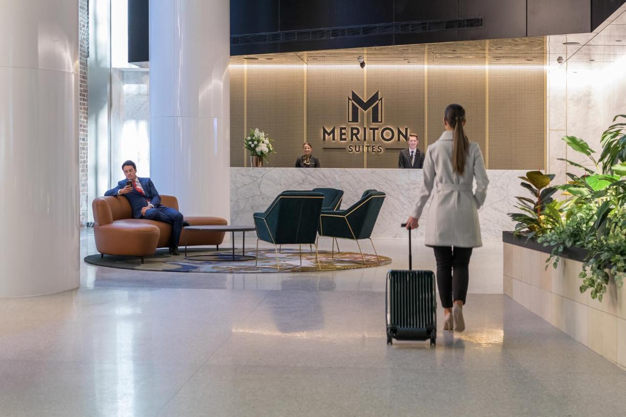Meriton Suites Sussex Street, Sydney Exteriér fotografie The photo depicts a modern hotel lobby. In the foreground, a woman is walking towards the reception desk, pulling a suitcase behind her. She is dressed in a light-colored coat. To her left, there’s a comfortable seating area with a person sitting on 