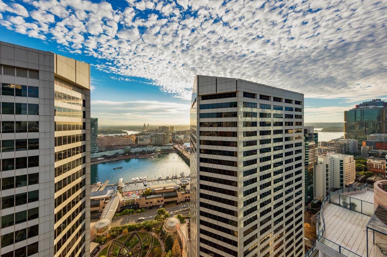 Meriton Suites Sussex Street, Sydney Exteriér fotografie The photo depicts a panoramic view of a modern cityscape featuring tall commercial buildings with glass facades. The shot is taken from a height, showing the intricate architecture of the nearby structures. Below, there is a body of water, which appe