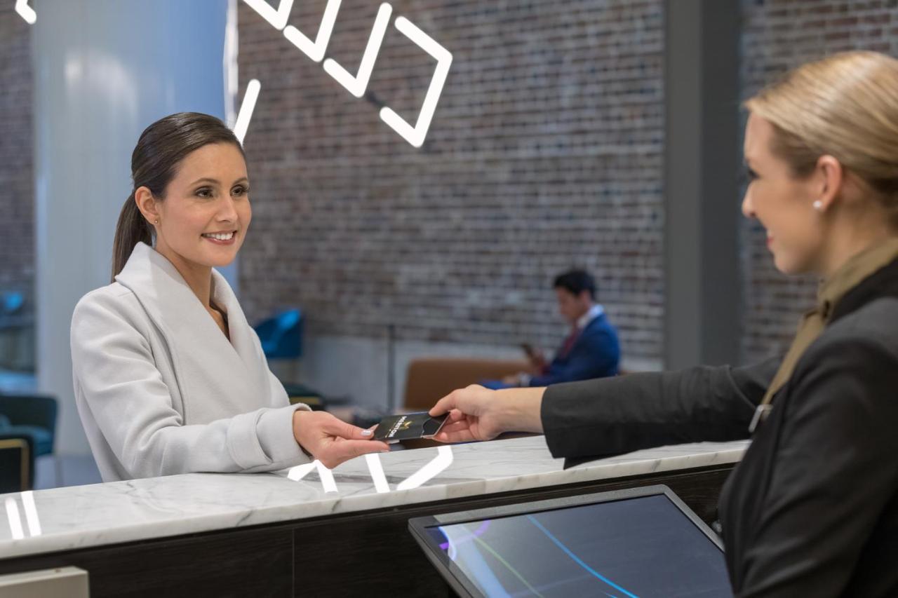 Meriton Suites Sussex Street, Sydney Exteriér fotografie The image depicts a scene at a hotel reception desk. A woman with long hair, dressed in a white outfit, is smiling as she hands her card to a receptionist, who is in a black uniform. The reception area features a modern design, and there is a person 
