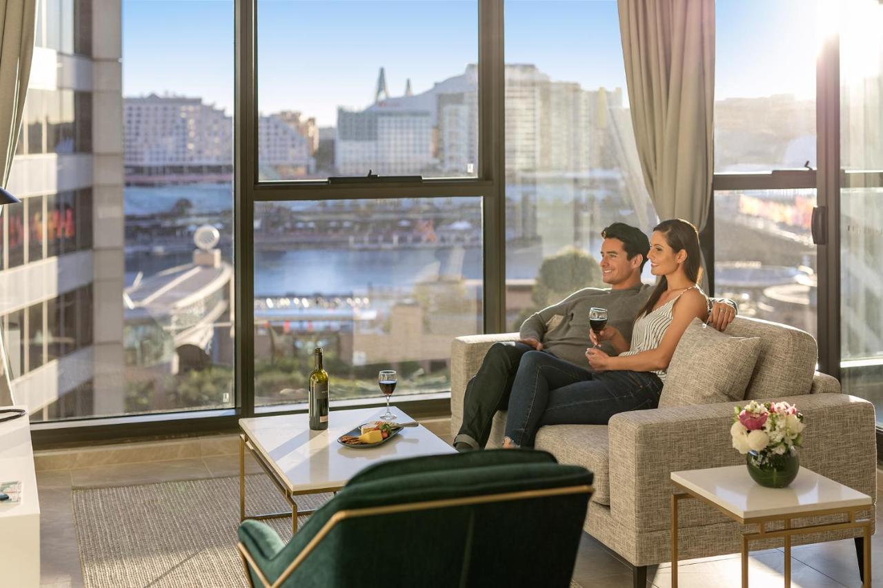 Meriton Suites Sussex Street, Sydney Exteriér fotografie The photo shows a cozy indoor setting with a couple sitting closely together on a light-colored sofa. They are holding drinks, likely wine, and appear to be enjoying each other's company while looking out of a large window. The window provides a view