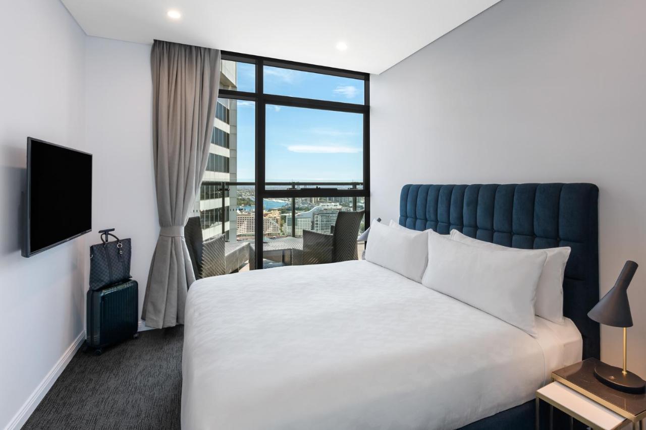 Meriton Suites Sussex Street, Sydney Exteriér fotografie The photo shows a modern hotel room featuring a double bed with white linens and a tufted blue headboard. There is a small bedside table next to the bed. A large window allows natural light to fill the room, offering a view of the city outside, inclu