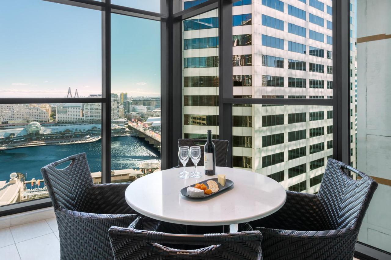 Meriton Suites Sussex Street, Sydney Exteriér fotografie The photo shows a modern dining area with a round table and four woven chairs. On the table, there are two wine glasses, a bottle of wine, and a plate with food items. The large windows provide a view of a cityscape featuring tall buildings and a bod