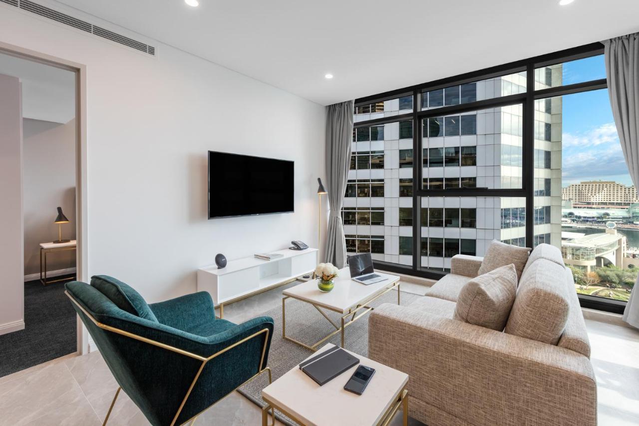 Meriton Suites Sussex Street, Sydney Exteriér fotografie The photo shows a modern living room with large windows that let in natural light, providing a view of a cityscape outside. The interior features a comfortable light-colored sofa and a stylish dark green armchair. A sleek TV is mounted on the wall op