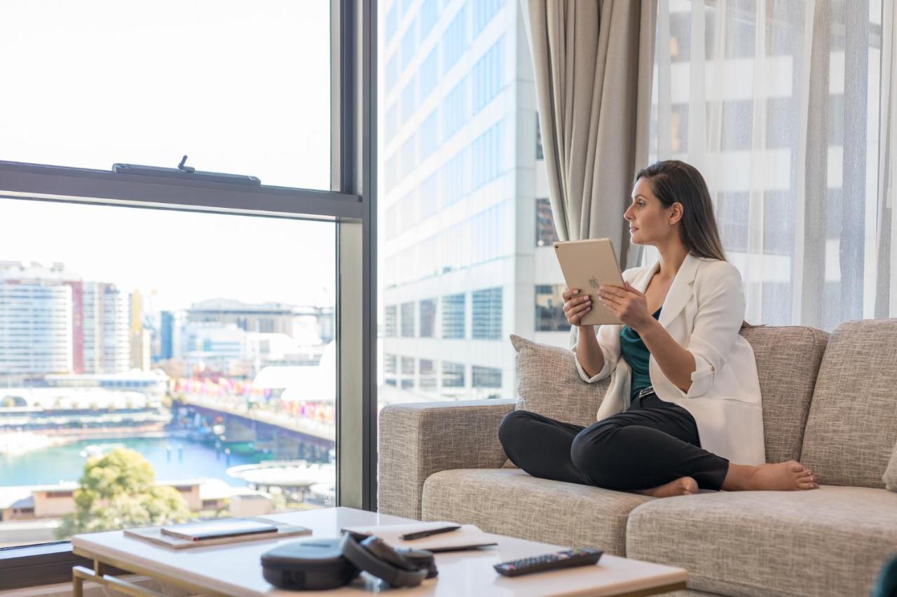 Meriton Suites Sussex Street, Sydney Exteriér fotografie The image portrays a woman sitting cross-legged on a light-colored sofa, engaged with a tablet in her hands. She is dressed in a white blazer and dark pants, appearing relaxed and focused. The setting features large windows that provide a view of a w