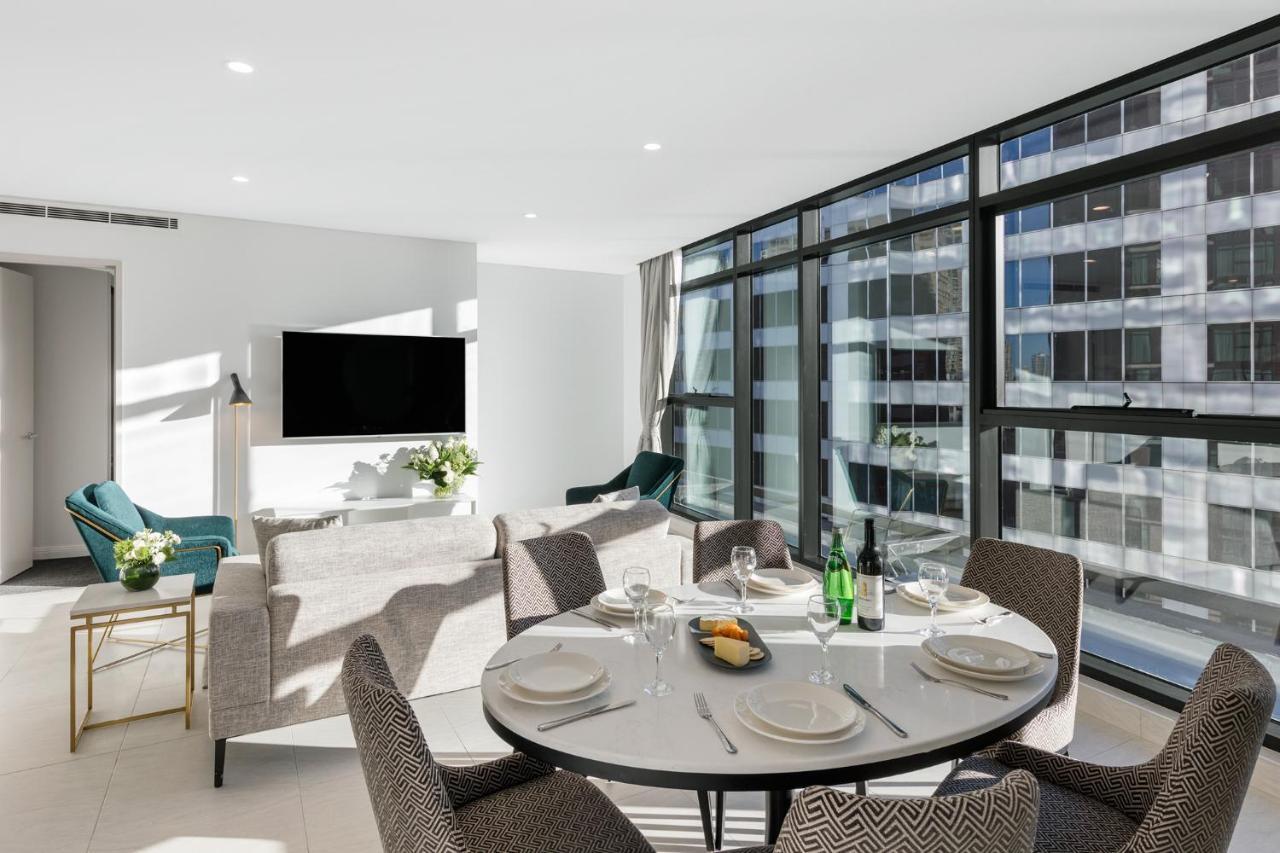 Meriton Suites Sussex Street, Sydney Exteriér fotografie The photo shows a modern and stylish living room and dining area. In the foreground, there is a round dining table set for a meal with plates, cutlery, and drinks. The seating arrangement includes comfortable chairs around the table. 

In the backgro
