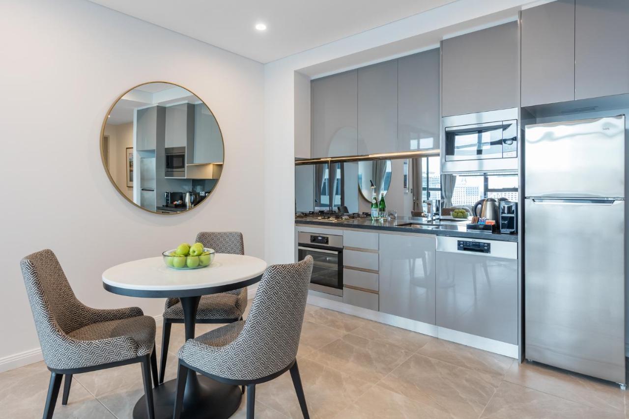 Meriton Suites Sussex Street, Sydney Exteriér fotografie The photo features a modern kitchen and dining area. The kitchen has sleek grey cabinets and countertops, with built-in appliances including an oven and possibly a cooktop. There's a large stainless steel refrigerator on the right. A round dining tab