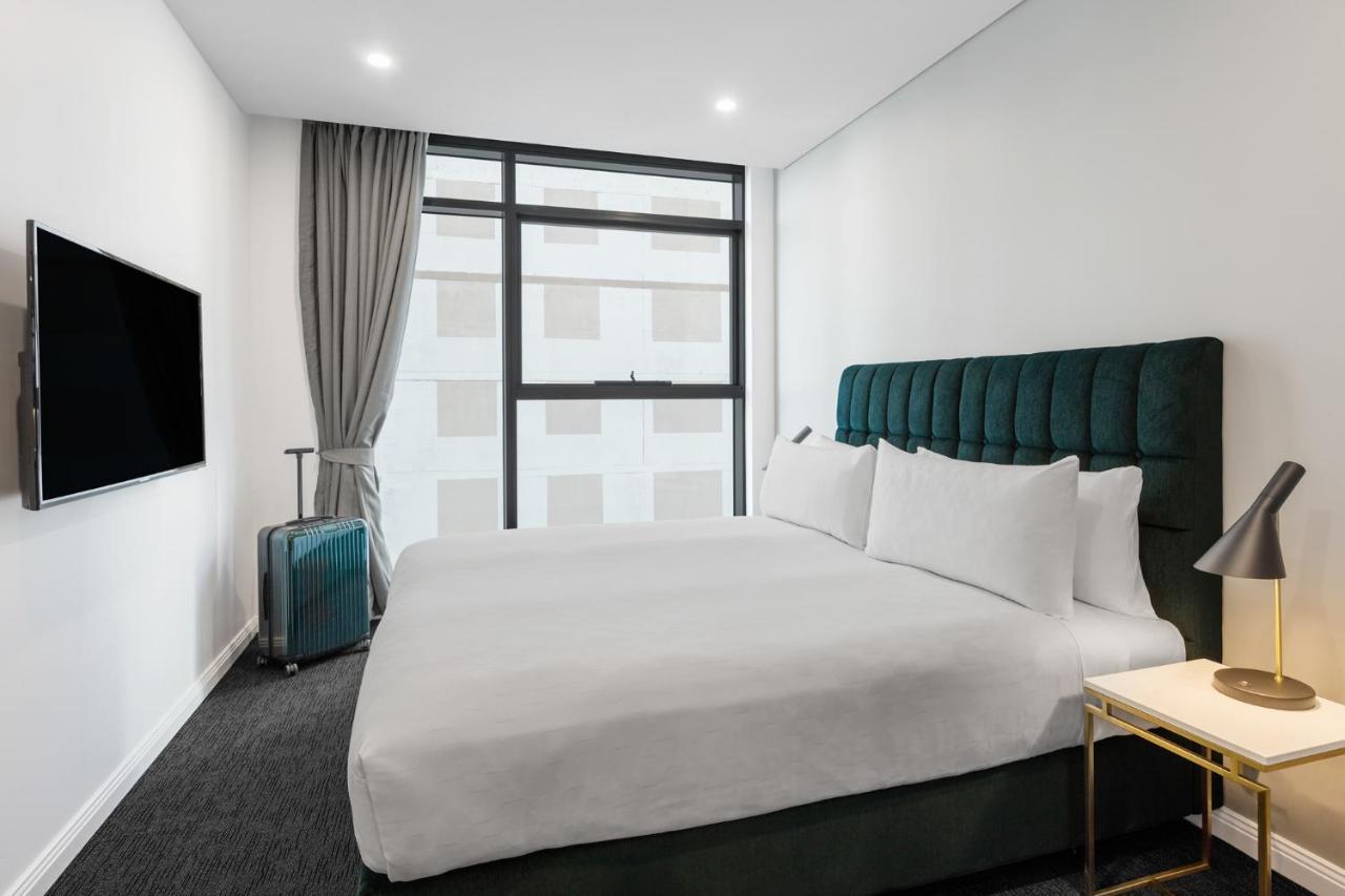 Meriton Suites Sussex Street, Sydney Exteriér fotografie The photo shows a modern hotel room. It features a large bed with a dark green tufted headboard and multiple white pillows. There's a nightstand with a lamp beside the bed. A sleek television is mounted on the wall opposite the bed, and a suitcase is