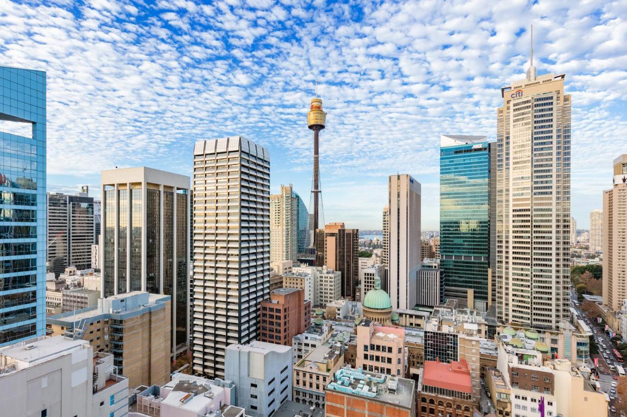 Meriton Suites Sussex Street, Sydney Exteriér fotografie The photo shows a panoramic view of a city skyline dominated by tall skyscrapers. In the center, there is a notable tower with a distinctive golden structure at its top, which is likely a landmark of the city. The buildings have a mix of modern glass