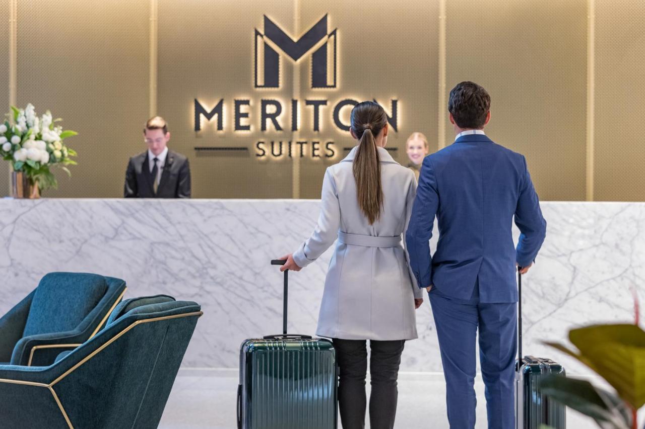Meriton Suites Sussex Street, Sydney Exteriér fotografie The photo shows a couple standing at a reception area in a hotel named "Meriton Suites." They are both facing the reception desk, where staff members are visible. The man is dressed in a suit, and the woman is wearing a light-colored coat. They each 