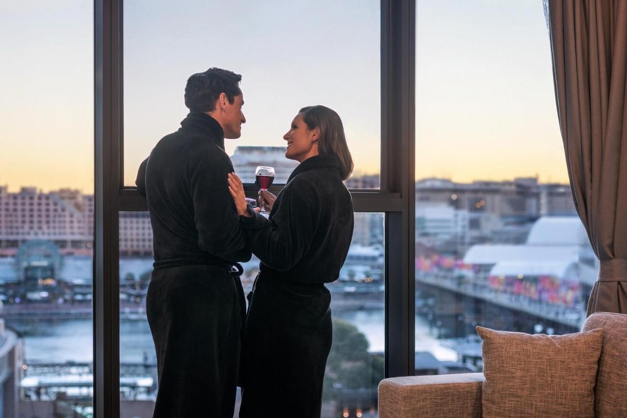 Meriton Suites Sussex Street, Sydney Exteriér fotografie The photo depicts a couple standing closely together by a large window. They are wearing dark bathrobes and holding drinks, gazing into each other’s eyes. The background features a scenic view, likely of a cityscape at sunset, with soft light illumin