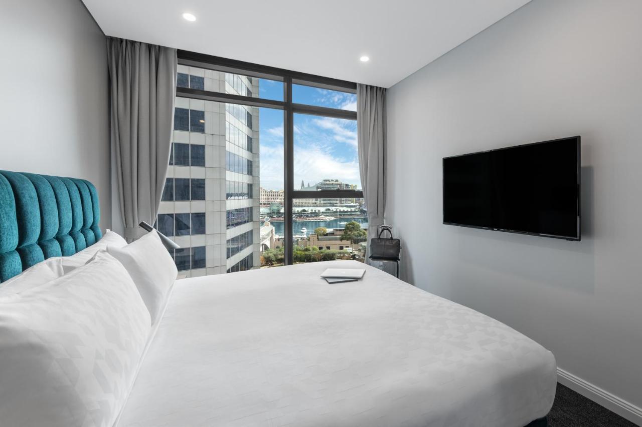 Meriton Suites Sussex Street, Sydney Exteriér fotografie The photo shows a modern hotel room with a large bed featuring a white comforter and pillows. There is a blue headboard that adds a pop of color to the decor. A large window provides a view of the outside, which includes buildings and a glimpse of wa