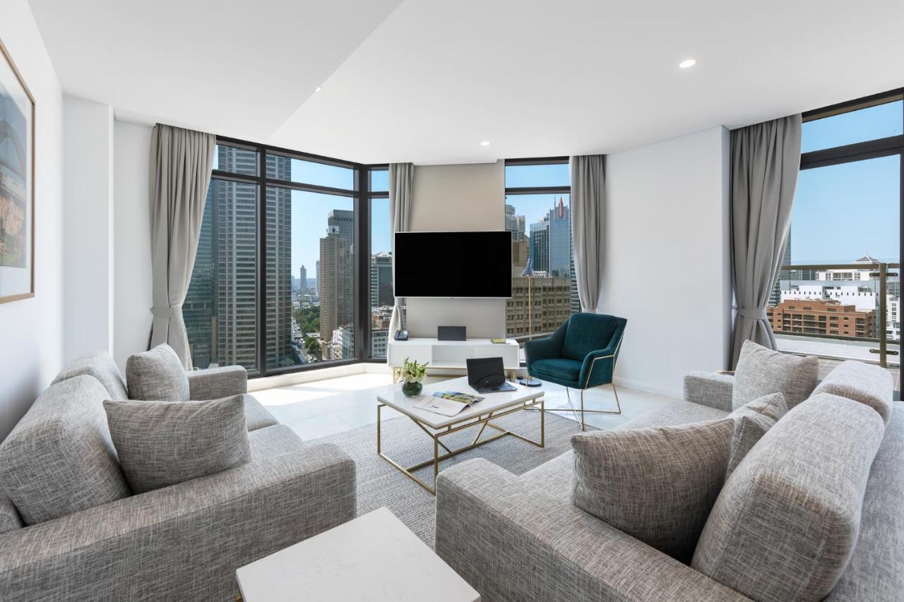 Meriton Suites Sussex Street, Sydney Exteriér fotografie The photo showcases a modern living room with large windows offering a view of a city skyline. The room features a light color palette with gray sofas arranged around a coffee table. A flat-screen TV is mounted on the wall, and there is a green armch