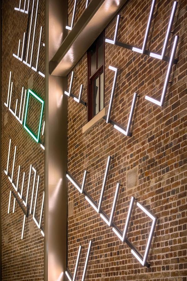 Meriton Suites Sussex Street, Sydney Exteriér fotografie The photo shows a brick wall featuring a design made up of linear light fixtures. These light strips create geometric shapes and patterns, with some sections illuminated in white and one section highlighted in green. The combination of the brick text