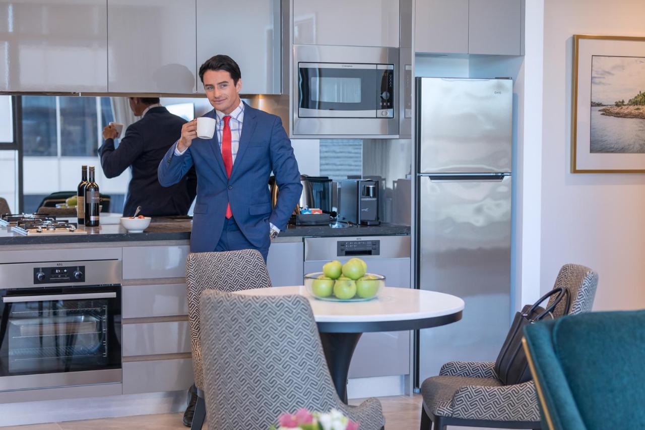 Meriton Suites Sussex Street, Sydney Exteriér fotografie The photo depicts a well-dressed man in a suit standing in a modern kitchen. He is holding a cup, possibly of coffee, and appears to be looking towards the camera. The kitchen features contemporary appliances, including a microwave and refrigerator. 