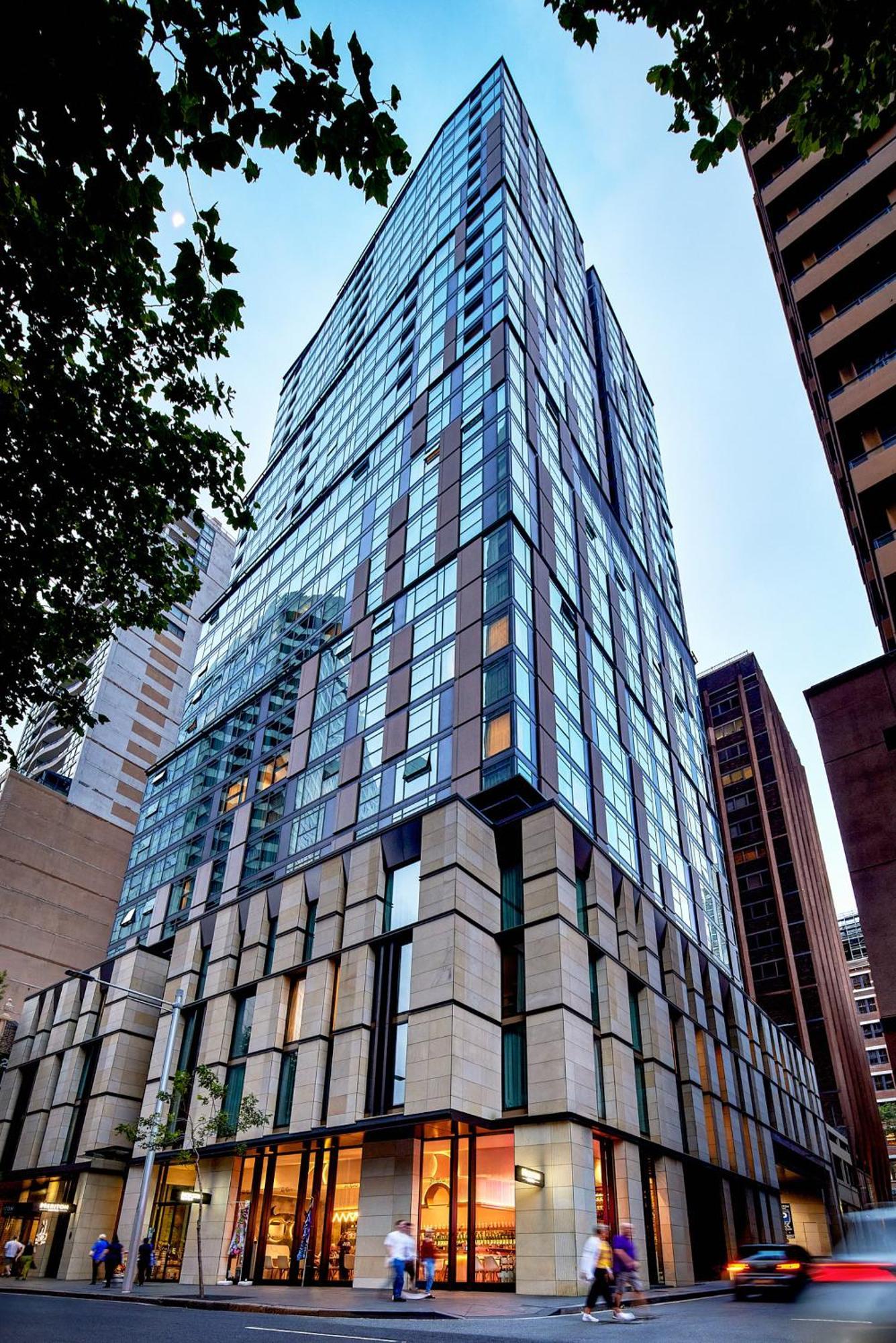 Meriton Suites Sussex Street, Sydney Exteriér fotografie The photo shows a modern high-rise building with a sleek, glass facade reflecting its surroundings. The structure features a combination of glass and concrete materials, with multiple stories visible. The lower part of the building has a sturdy, text