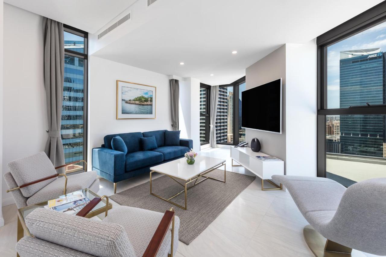 Meriton Suites Sussex Street, Sydney Exteriér fotografie The photo shows a modern living room featuring large windows that allow plenty of natural light. The room is elegantly designed with a blue sofa, a light-colored coffee table, and two armchairs. There’s a flat-screen TV mounted on the wall, and a pie